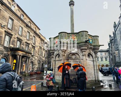 Edinburgh, Schottland, Vereinigtes Königreich - 15. Februar 2026: Touristen versammeln sich unter orangefarbenen Regenschirmen um das verzierte Mercat Cross im Zentrum von Edinburgh während eines nassen, Stockfoto