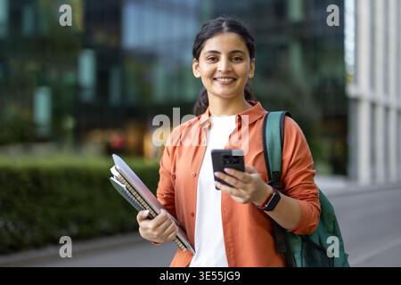 Glückliche junge indianerin in legeren Kleidern mit Rucksack und Notizbüchern. Sie lächelte in die Kamera, während sie ihr Handy auf einem modernen Universitätsgelände überprüft Stockfoto