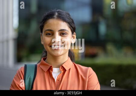 Die junge indische Studentin lächelt selbstbewusst und blickt auf die Kamera, während sie draußen auf einem Universitätscampus steht. Sie reflektiert Bildung, Jugend und zukünftigen Erfolg Stockfoto