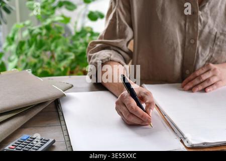 Frau sitzt am Schreibtisch mit Aktenmappen und Papierkram und schreibt mit Stift auf leeres Papier. Home-Office-Szene mit Schwerpunkt Organisation, Dokumentation und Stockfoto
