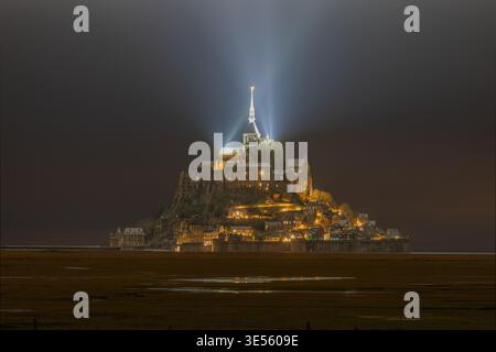Blick auf die beleuchtete Abtei Mont Saint-Michel bei Nacht mit einem hellen Scheinwerferstrahl vom Turm über die Wattflächen in Mont Saint-Michel, Norman Stockfoto
