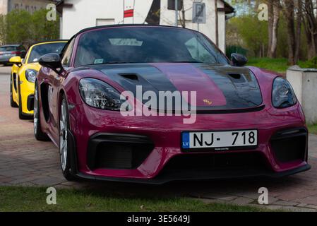 Valmiera, Lettland-26.2025-zwei Porsche Sportwagen, ein Porsche 718 Cayman GT4 RS in Magenta und Schwarz und ein gelber Porsche, parkten draußen auf einem Backstein Stockfoto