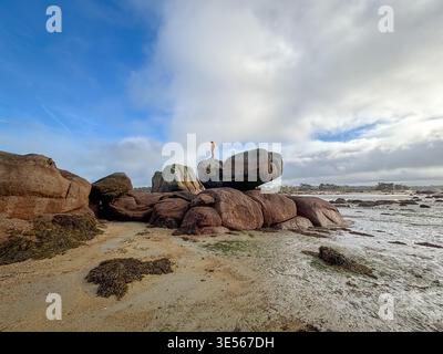Ein einsamer Mensch steht auf einem großen, ausgleichenden Felsen in Tregastel an der Pink Granit Coast in der Bretagne, Frankreich, umgeben von verstreuten Felsblöcken und nassem Sand Stockfoto