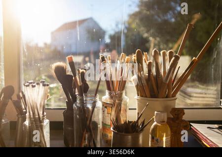 Sammlung von Pinseln und Kunstwerkzeugen, die in Gläsern auf einer Fensterbank aufbewahrt werden und durch natürliches Licht aus einem Fenster beleuchtet werden. Stockfoto