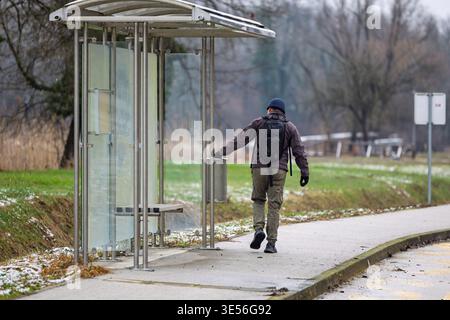 Mann mit Rucksack und Winterjacke geht an einem bewölkten, kühlen Tag vorbei an einer Glasbushaltestelle, die von hinten auf einem ruhigen Bürgersteig gefangen wird Stockfoto