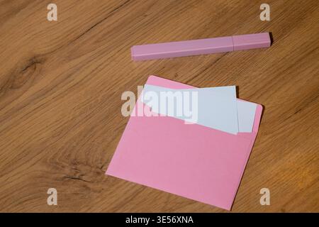 Ein rosafarbener Umschlag mit weißem Papier sitzt auf einem Holztisch neben einem rosafarbenen Stift, mit einem warmen und gemütlichen Hintergrund Stockfoto
