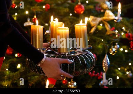 Hände halten einen Adventskranz mit vier Kerzen, hinter dem der Weihnachtsbaum sanft verschwimmt. Stockfoto