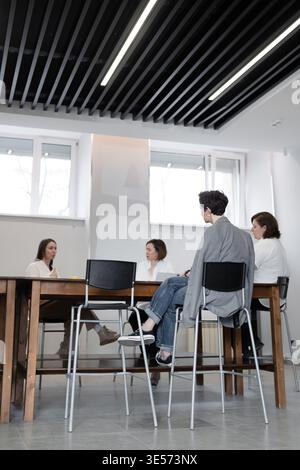 Das Business-Team sitzt im modernen Büro am Meeting-Tisch unter linearen Deckenleuchten und spricht. Team, Meeting, modern, Büro, sitzen, Diskussion. Stockfoto