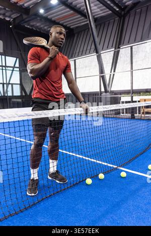Afroamerikaner in Sportbekleidung, der Schläger hält und sich mit gelben Bällen auf das Netz stützt Stockfoto