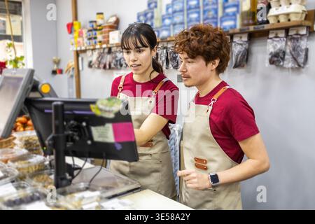Zwei Mitarbeiter in Lebensmittelgeschäften mit Schürzen stehen hinter dem Tresen und nutzen ein Point-of-Sale-System. Regale mit Waren sind in sichtbar Stockfoto