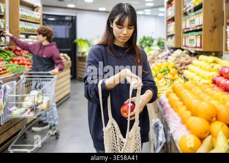 Eine Frau kauft in einem Lebensmittelgeschäft ein, wählt frische Äpfel aus und verstaut sie in ihre umweltfreundliche Tasche. Der Laden ist voll mit bunten Produkten Stockfoto