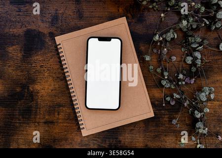Smartphone mit leerem Bildschirm auf Notebook mit Pflanze auf Holztisch, minimaler Arbeitsbereich flach liegend mit Kopierraum. Draufsicht Stockfoto