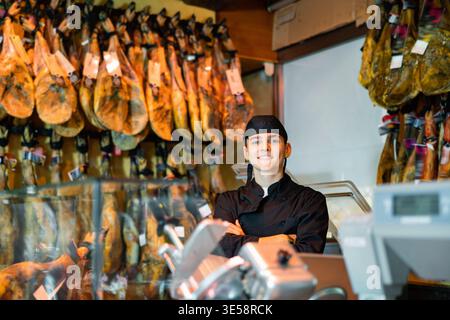 Junger Metzgerverkäufer in schwarzer Uniform, mit gekreuzten Armen in der Nähe eines Regals, mit hängenden Jamonbeinen Stockfoto