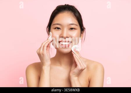 Frau, die Hautpflege auf ihrem Gesicht vor rosa Hintergrund in einer Beauty-Routine aufträgt Stockfoto