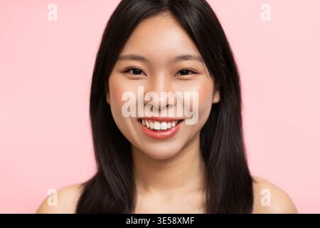 Lächelnde junge Frau mit langen dunklen Haaren steht vor einem rosafarbenen Hintergrund Stockfoto