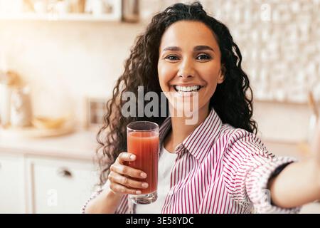 Die Frau trinkt und lächelt, während sie tagsüber Selfie in einer hellen Küche nimmt Stockfoto