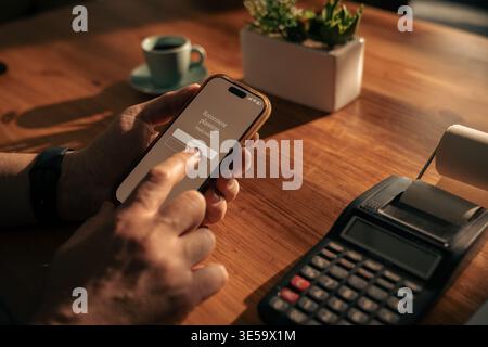 Reifer Mann, der zu Hause Smartphone mit mobiler Banking-Anwendung nutzt, persönliche Finanzen und digitale Zahlungen mit Taschenrechner auf einem Holztisch verwaltet. Stockfoto