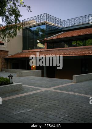 Leerer gepflasterter Innenhof, umgeben von einem renovierten Gebäude mit traditionellen roten Ziegeldächern und einem modernen Glasobergeschoss in der Dämmerung. Th Stockfoto