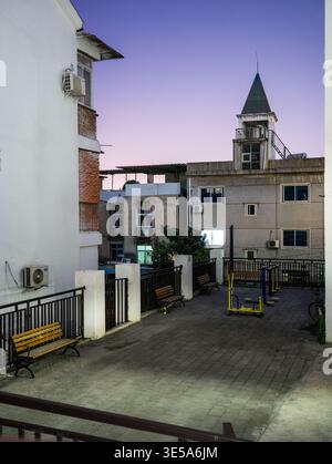 Leerer gepflasterter Innenhof in einem Wohnviertel mit Holzbänken, Fitnessgeräten im Freien und einem kleinen Turmgebäude in der Dämmerung. Die s Stockfoto