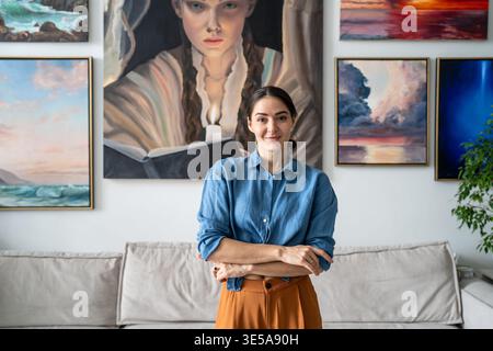 Erfolgreiche Malerin posiert vor der Kamera mit selbstbewusst lächelndem Ausdruck vor eigenen Kunstwerken Stockfoto