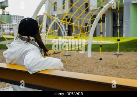 Die junge Frau in einer weißen Jacke sitzt leise auf einer Bank und blickt auf einen modernen Spielplatz mit heller Kletterausrüstung. Stockfoto