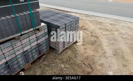 Gestapelte graue Betonpflastersteine, die mit grünen Bändern auf Holzpaletten neben einem Kiesbereich gesichert sind Stockfoto