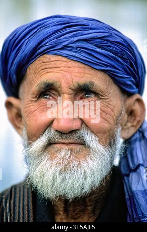 Ein alter Mann mit weißem Bart und blauem Turban lächelt. Er ist in Chiwa, Usbekistan, und beobachtet tagsüber seine Umgebung. Stockfoto