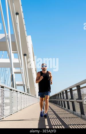 Städtischer Sportler, der auf einer modernen Stadtbrücke joggt: Vorderansicht eines Mannes, der im Freien aktiv ist. Stockfoto