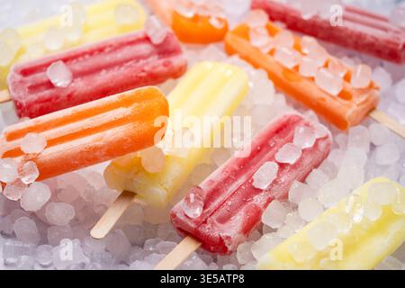 Nahaufnahme von farbenfrohem Fruchteis auf einem Bett aus zerstoßenem Eis, mit sichtbarem Frost und schmelzender Textur. Leuchtend gelbe, rote und orangene Stiele erzeugen einen Stockfoto
