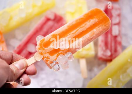 Nahaufnahme von farbenfrohem Fruchteis auf einem Bett aus zerstoßenem Eis, mit sichtbarem Frost und schmelzender Textur. Leuchtend gelbe, rote und orangene Stiele erzeugen einen Stockfoto