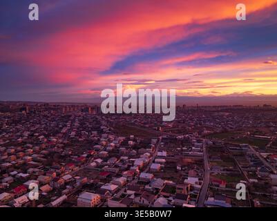 Aus der Vogelperspektive lässt sich die Zersiedelung der Stadt unter dem dramatischen Sonnenuntergangshimmel erfassen. Leuchtende rosa und orange Wolken stehen im Kontrast zu grauen Dächern. Keine Menschen sichtbar, außer sce Stockfoto