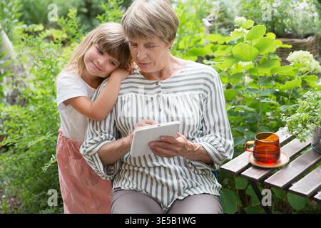 Großmutter und kleine Enkelin benutzen Tablet und haben gemeinsam Spaß im Garten. Glückliche Familie, die den Sommer im Freien genießt. Stockfoto