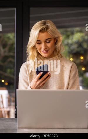Frau, die am Kaffeetisch sitzt und einen beigefarbenen Strickpullover mit Smartphone trägt und am Laptop arbeitet. Gemütlich, Glas, Grün, Bokeh, Fernbedienung, Coworking, Ambiente Stockfoto