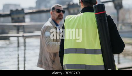Bauarbeiter in Warnweste spricht mit Kollegen am Wasser mit Blaudruckrohr Stockfoto