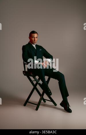 Ein gutaussehender Mann in einem maßgeschneiderten Anzug sitzt elegant auf einem Direktorenstuhl und zeigt Stil. Stockfoto