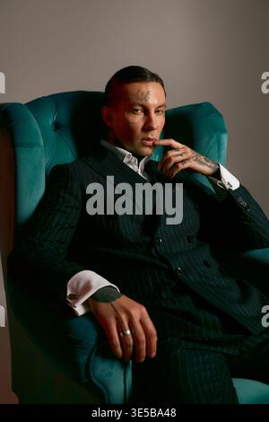 Ein junger Mann mit auffälligen Details sitzt elegant in einem schicken Stuhl und strahlt moderne Mode aus. Stockfoto
