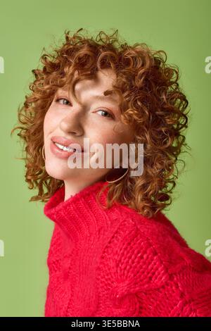 Eine fröhliche junge Frau mit lockigen roten Haaren lächelt hell, während sie einen kuscheligen roten Pullover trägt. Stockfoto