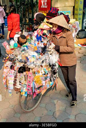 Ein Verkäufer schiebt einen Fahrradwagen voller bunter Gegenstände in Hanoi. Leute gehen vorbei, während sie ihre Waren in der geschäftigen Straßenlage verkauft. Stockfoto