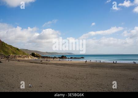 Looe Beach, Cornwall, England – Menschen, die an einem hellen Frühlingstag entlang der Sandy Shore mit Green Hills und Blue Sea spazieren Stockfoto