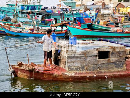 Ein Mann steht auf einem hölzernen Fischerboot in Phu Quoc, Vietnam. Nahe gelegene Boote liegen an und das Wasser reflektiert das Sonnenlicht. Stockfoto