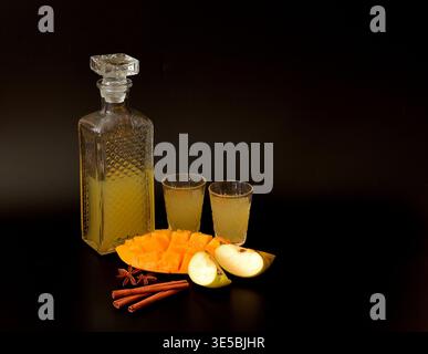 Obstlikör mit Gewürzen in Kristallglas auf schwarzem Hintergrund, neben reifer Mango und grünem Apfel. Nahaufnahme. Stockfoto