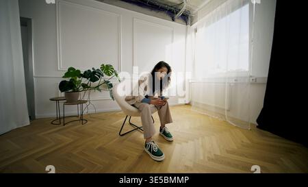 Stilvolle junge Frau in lässiger Mode, die in modernem minimalistischem Interieur sitzt Stockfoto