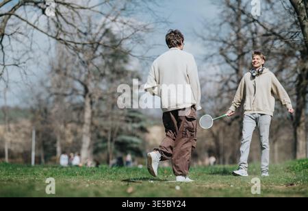 Zwei Freunde spielen Badminton und lachen gemeinsam in einem sonnigen Stadtpark an einem Frühlingstag Stockfoto