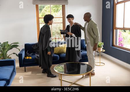 Verschiedene männliche Familien feiern mit schwarzen Abschlusskleidern und Mortarboards im Wohnzimmer Stockfoto