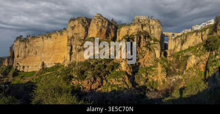 Ronda ist in dieser Szene mit Klippen und Gebäuden zu sehen. Die Aussicht zeigt die berühmte Brücke der Stadt. Dunkle Wolken füllen den Himmel, während die Sonne über dem Land untergeht Stockfoto