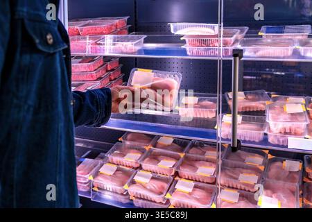 Lebensmittelhändler wählen Fleischprodukte aus. Supermarktkunde wählt Rindfleisch unter hellen Lichtern. Der Käufer untersucht verpacktes Fleisch im Kühlschrank Stockfoto