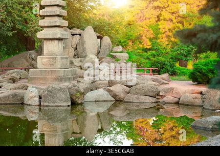 Eine ruhige japanische Gartenszene mit einer Steinpagode umgeben von großen Felsen und einem Teich, der die Landschaft widerspiegelt, mit lebhaftem Grün und Herbst Stockfoto