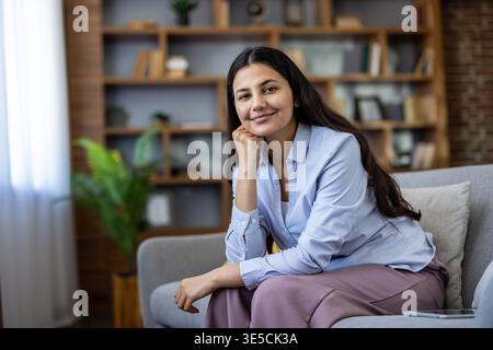Junge indianerin sitzt auf einem bequemen Sofa in ihrem modernen Wohnzimmer, lächelt in die Kamera und findet in der Freizeit zu Hause Komfort und Entspannung Stockfoto