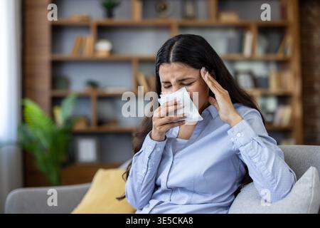 Junge Frau, die sich zu Hause unwohl fühlt, Kopfschmerzen und Niesen in ein Gewebe erlebt, symbolisiert Erkältung, Grippe, Virus oder Allergie-Symptome, während sie versucht, sich zu entspannen Stockfoto