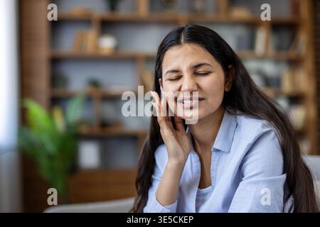 Junge Frau, die ihre Augen schließt und ein schmerzhaftes Ohr berührt, vor Ohrenschmerzen oder Tinnitus zuzwinkernd, Unwohlsein und Erschöpfung in einer offenen Innenumgebung zeigt Stockfoto